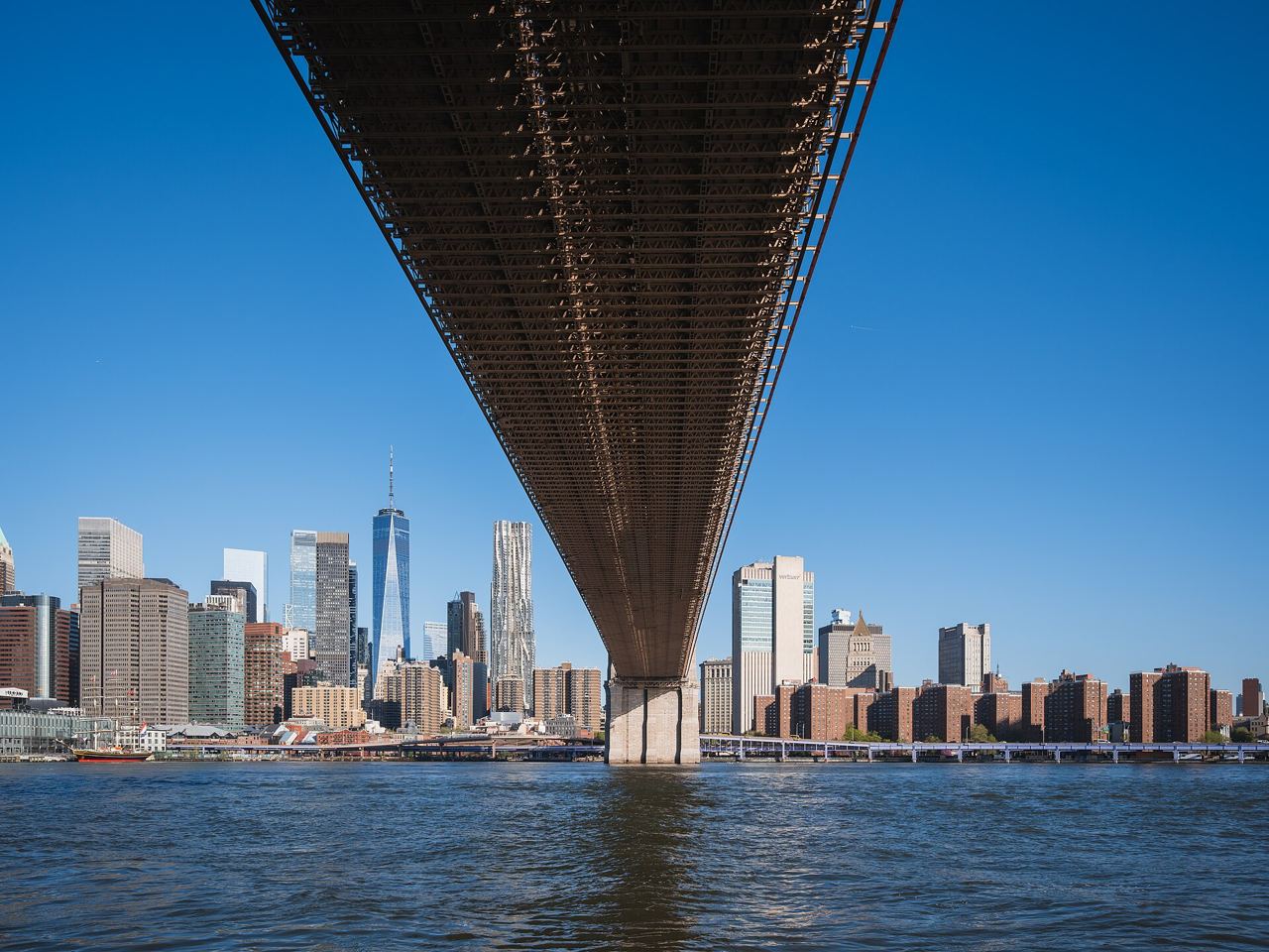 Pohled zespodu na ocelovou mostovku Brooklyn Bridge s mrakodrapy Dolního Manhattanu v pozadí
