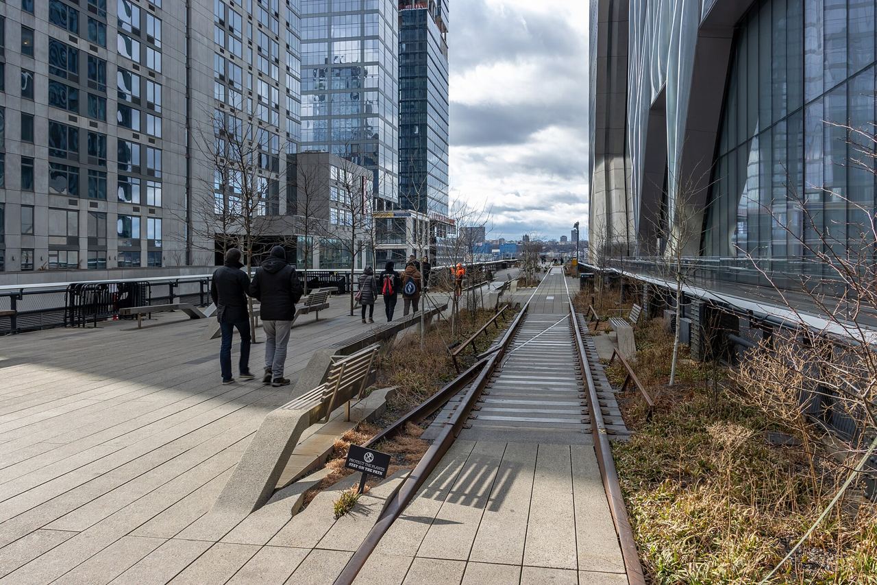 Dřevěná promenáda a původní koleje na High Line mezi moderními skleněnými budovami