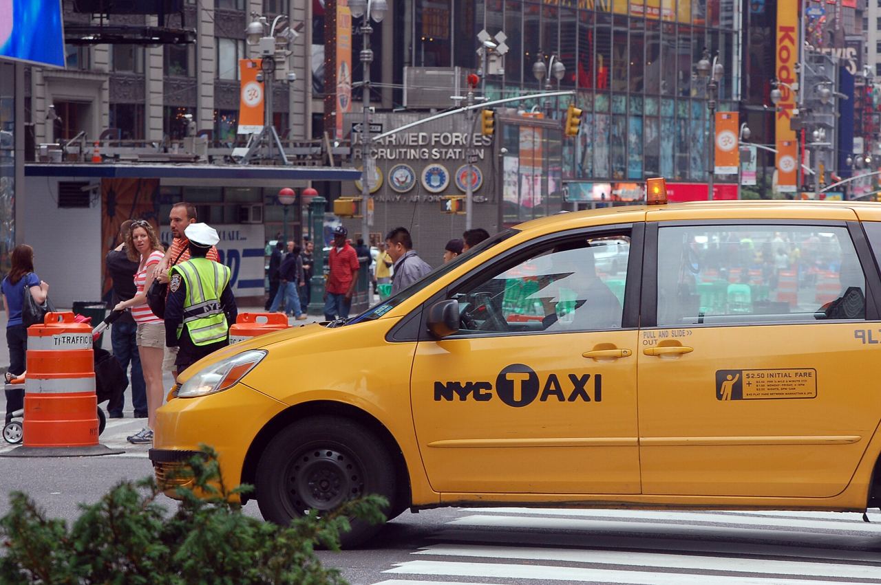Žluté taxi projíždí přes Times Square, v pozadí chodci a pracovníci u dopravních kuželů